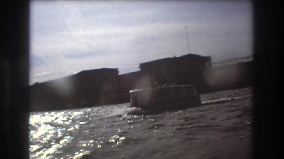 1975 Boston waterfront view from a moving boat, with another small craft crossing rippled water in front of dark shoreline buildings.