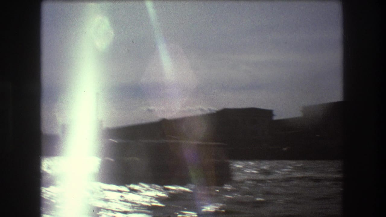 Flared 8mm frame from 1975 Boston harbor footage, showing water, a low shoreline structure, and a dark boat shape near the edge of the frame.