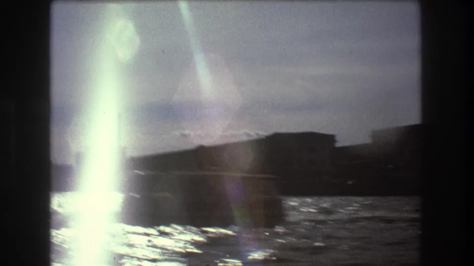 Flared 8mm frame from 1975 Boston harbor footage, showing water, a low shoreline structure, and a dark boat shape near the edge of the frame.