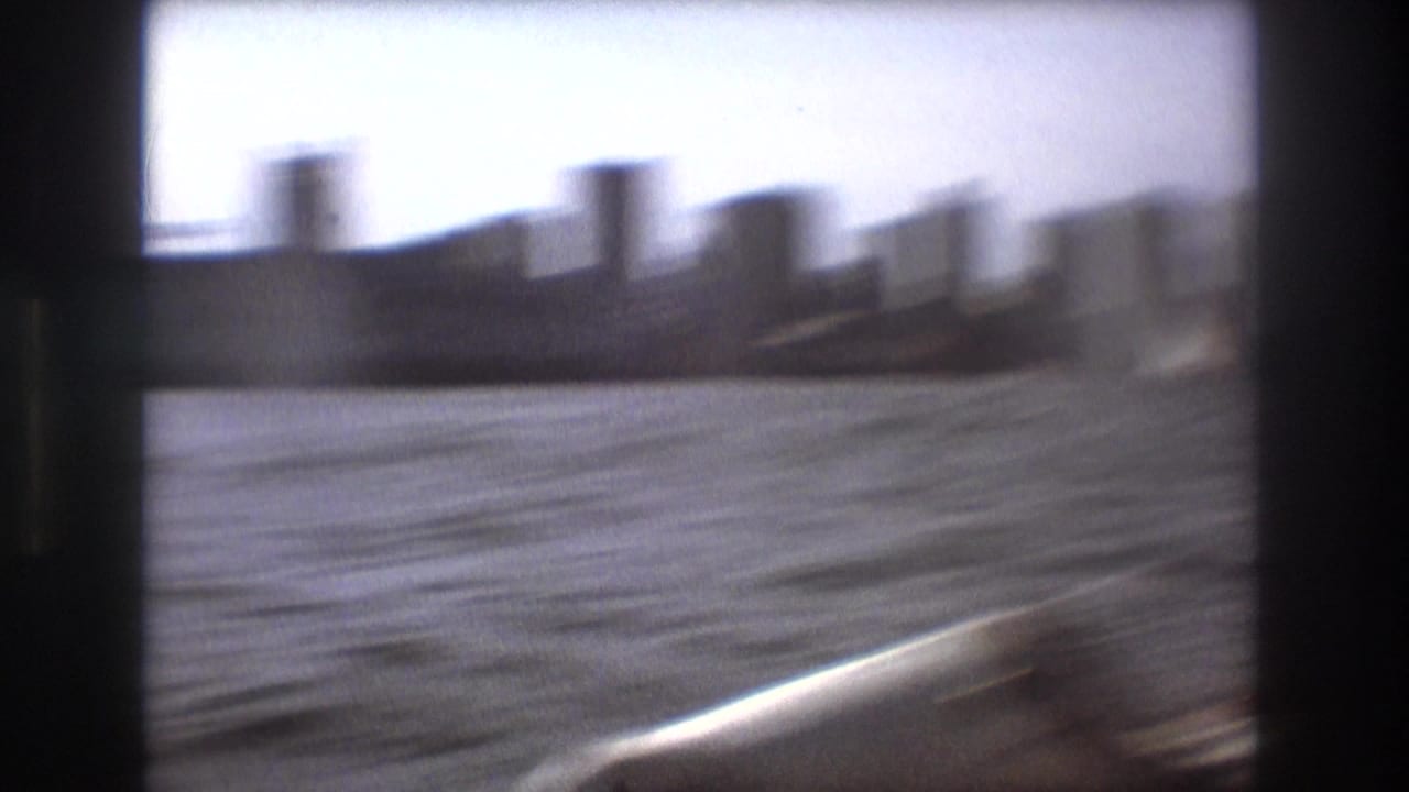 Soft, motion-blurred 1975 Boston harbor frame with distant high-rise forms, open water, and the edge of the filming boat in the foreground.