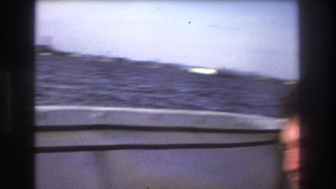 1975 Boston 8mm frame from a moving boat, showing choppy bay water, the boat edge, and a blurred passenger face at the lower right.