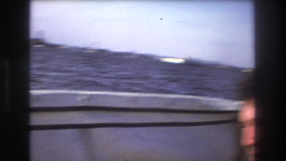 1975 Boston 8mm frame from a moving boat, showing choppy bay water, the boat edge, and a blurred passenger face at the lower right.