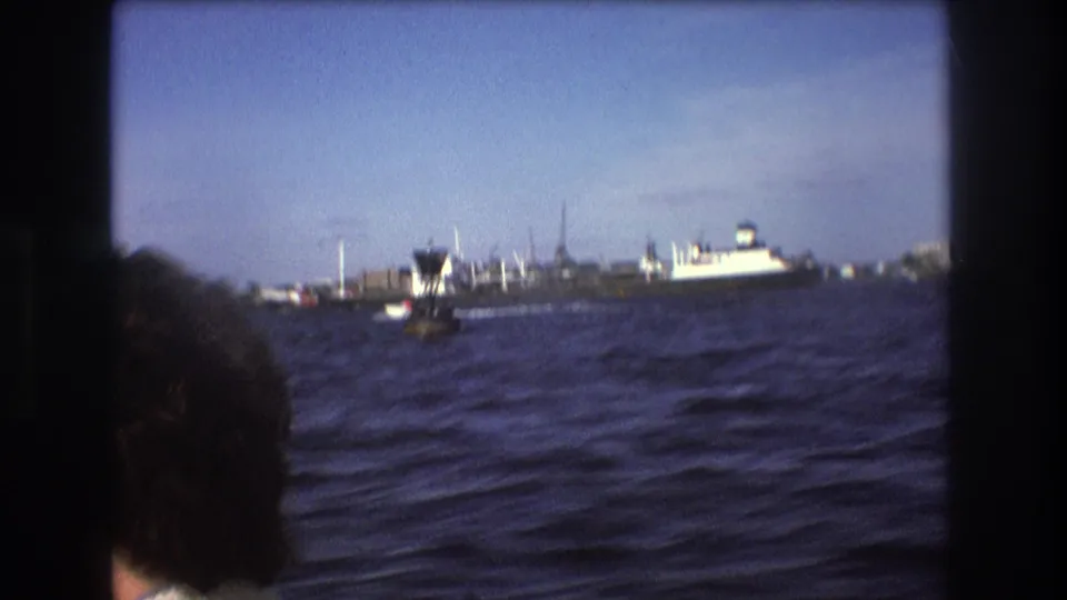 1975 Boston harbor view from a moving boat, with windblown passenger hair in the foreground and a large vessel and smaller boats in the distance.