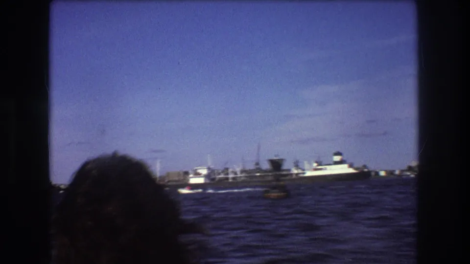1975 Boston 8mm boating frame showing a passenger's head at lower left, rough bay water, and a large ship near distant harbor structures.