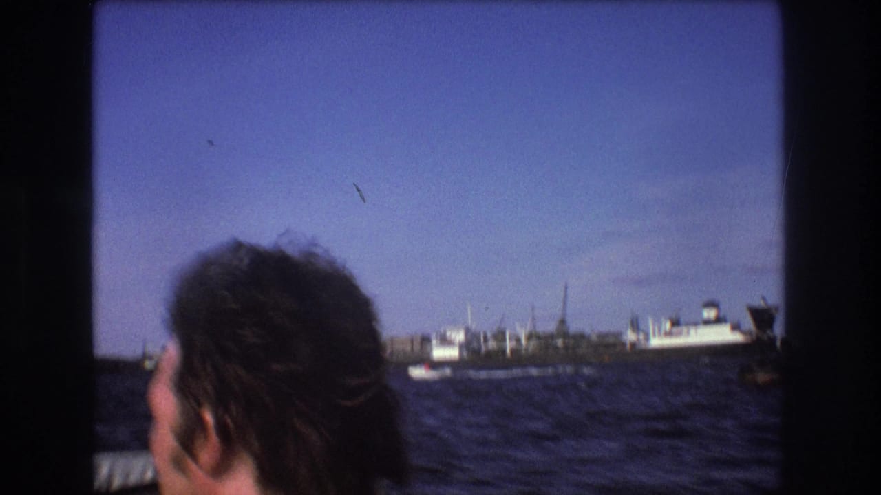 1975 Boston boat-ride frame with a passenger seen from behind, choppy water below, and ships and waterfront structures on the horizon.