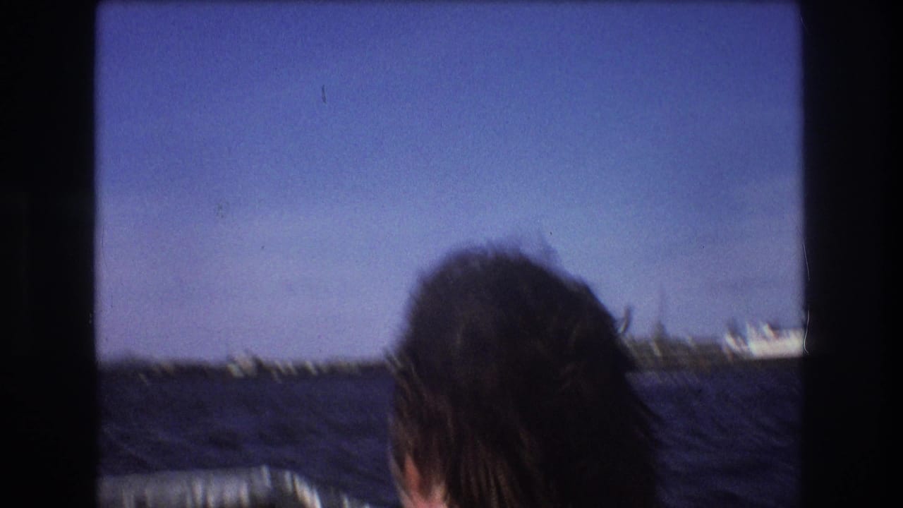1975 Boston home-movie frame with a man’s dark hair partly visible in the foreground as a small boat moves across blue harbor water toward a distant shoreline.