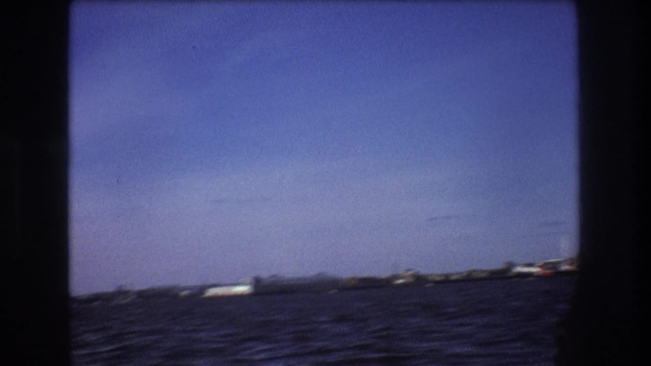 Open water seen from a moving boat with distant ships and shoreline under blue sky.