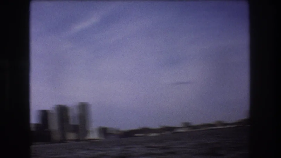A motion-softened 1975 Boston waterfront view shows choppy harbor water, low shoreline buildings, and distant vertical structures from a moving small boat.