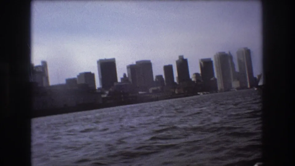 The Boston skyline appears across choppy harbor water in a slightly tilted 1975 8mm home-movie frame shot from a moving boat.