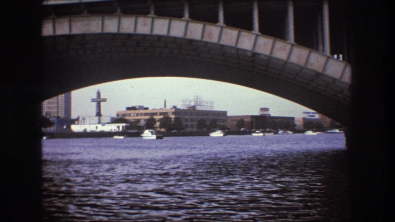 Boston-area 8mm boat view showing rippled water beneath a large arched bridge, with distant waterfront buildings and sky beyond.