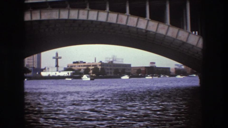 Boston-area 8mm boat view showing rippled water beneath a large arched bridge, with distant waterfront buildings and sky beyond.