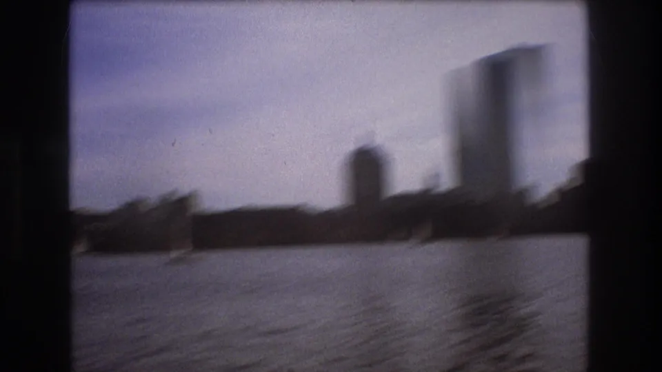 Soft-focus 8mm boat-view frame of Boston waterfront silhouettes and taller buildings across the water.