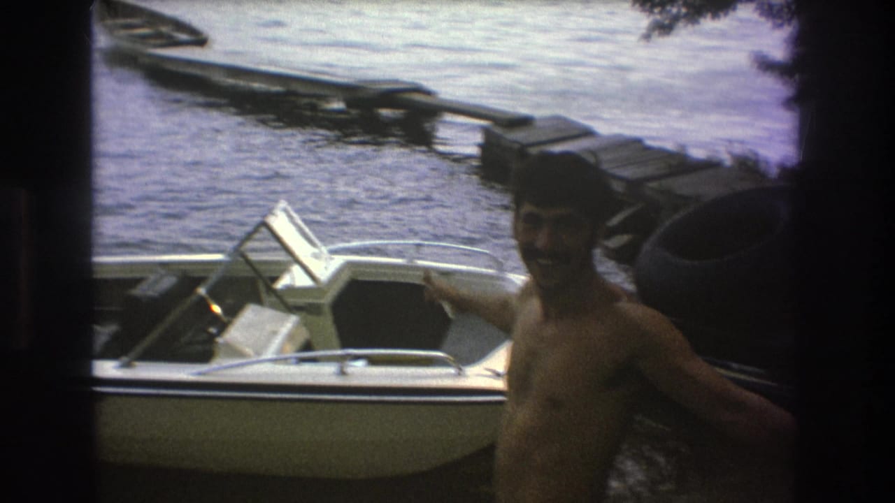 A shirtless adult man smiles near a white motorboat at a wooden lakeside dock in 1975 home-movie footage from the Boston metadata set.