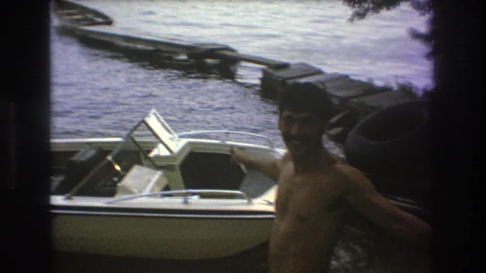 A shirtless adult man smiles near a white motorboat at a wooden lakeside dock in 1975 home-movie footage from the Boston metadata set.