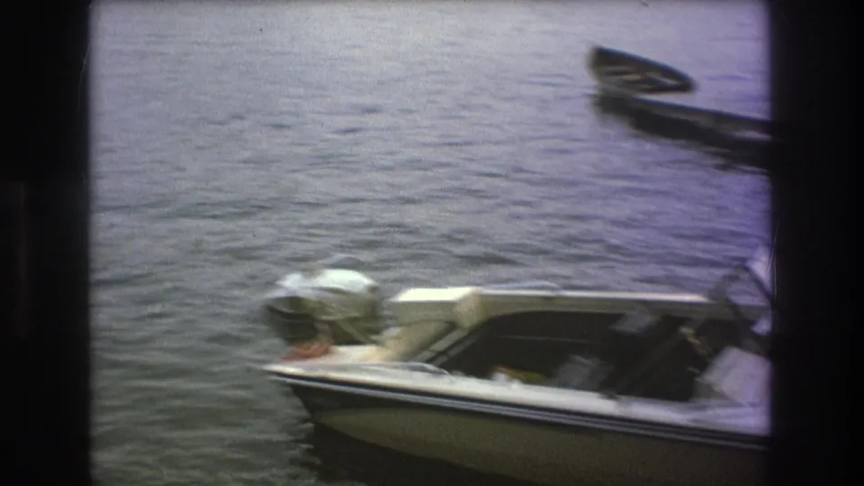 A white motorboat with an outboard motor sits on calm lake water near a dock, shown in soft 1970s 8mm home-movie footage.