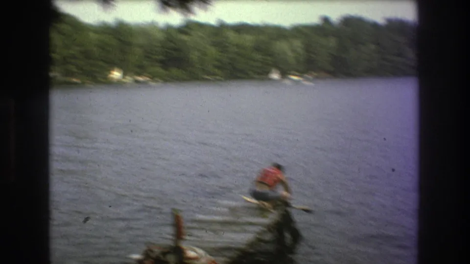 A distant water skier wearing a red life vest moves across the lake, with wooded shoreline visible in the background.