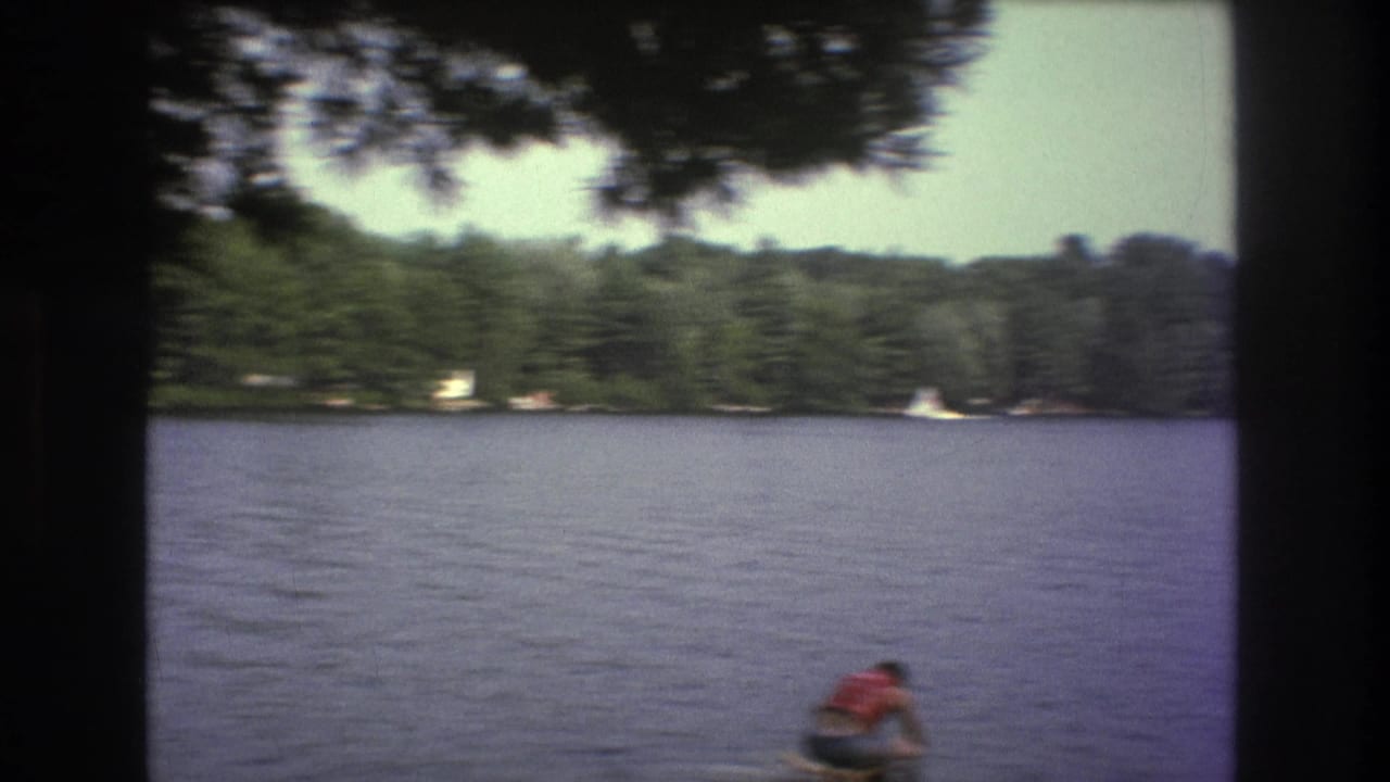 A wide 8mm lake view shows a distant water skier in a red life vest below a tree-lined shoreline, consistent with the supplied Boston 1975 clip metadata.