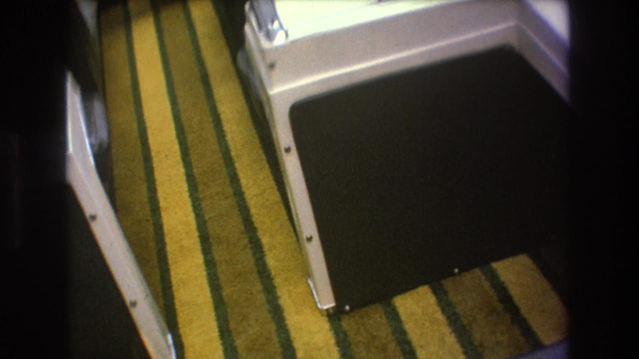 1975 8mm home-movie frame showing yellow and green striped carpeting inside a boat, bordered by white molded seating or cabinetry and a dark cushion area.
