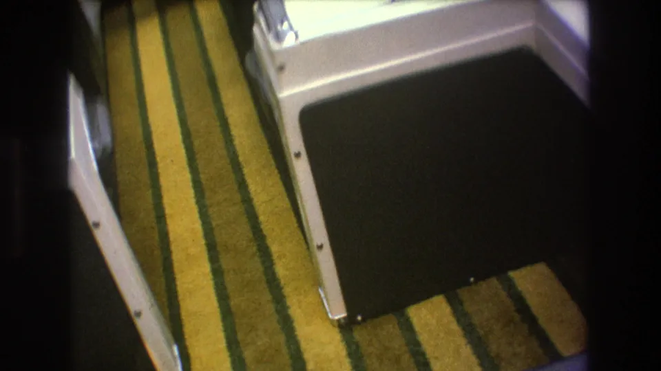 1975 8mm home-movie frame showing yellow and green striped carpeting inside a boat, bordered by white molded seating or cabinetry and a dark cushion area.