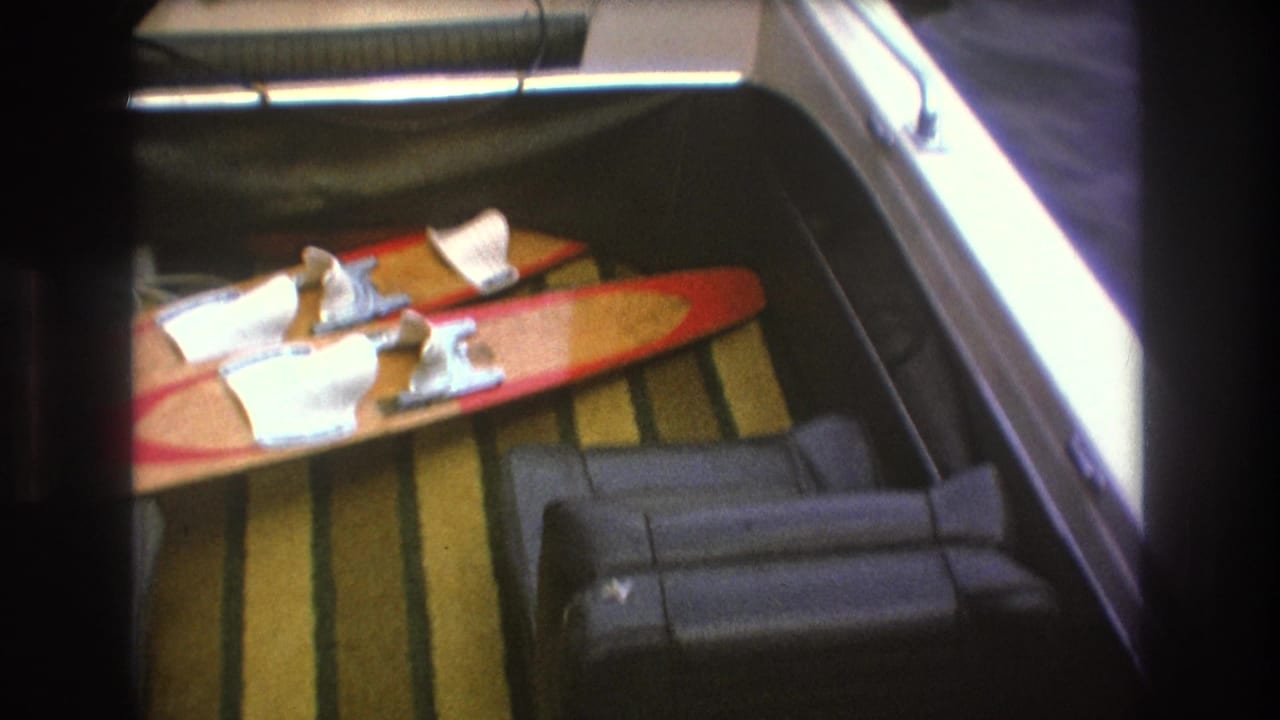 1975 Boston-area boating frame showing red water skis, papers or cloth, small gear, striped carpet, dark seating, and bright window light inside the vessel.