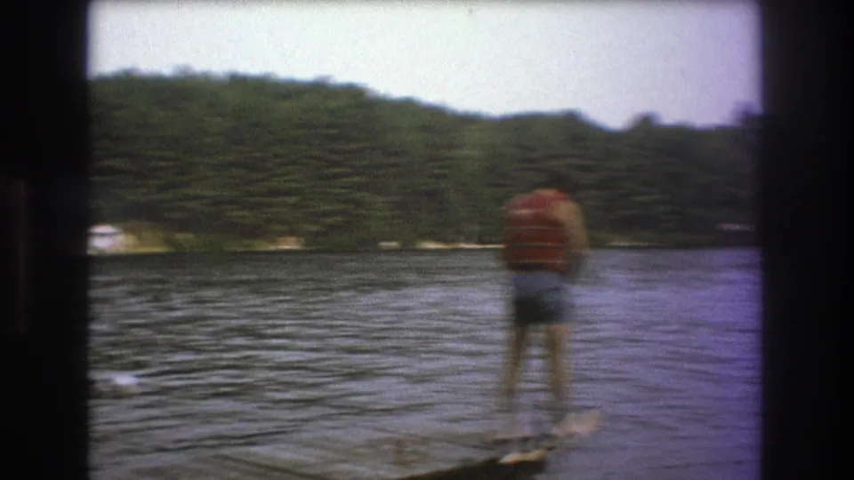 A skier in a red life vest and denim shorts stands on water skis on a lake, with a dense tree-lined shore in the background.