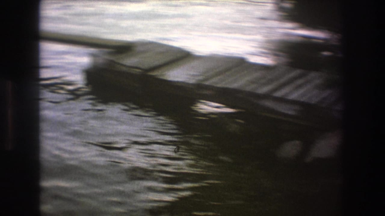 A dark wooden dock or low platform sits at the edge of rippling lake water, seen in a soft, handheld 8mm frame.