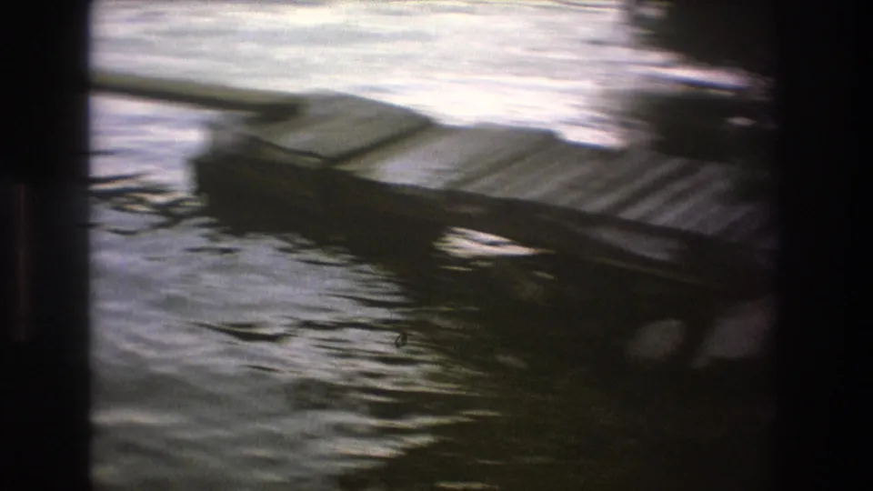 A dark wooden dock or low platform sits at the edge of rippling lake water, seen in a soft, handheld 8mm frame.