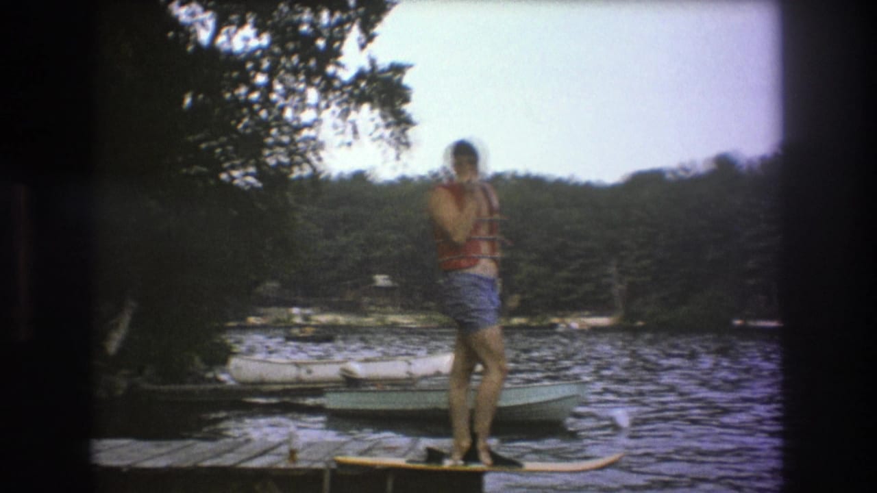 In metadata-supported 1975 Boston-area 8mm footage, a man in a red life vest and blue swim trunks stands on a wooden dock with lake water, boats, and trees behind him.