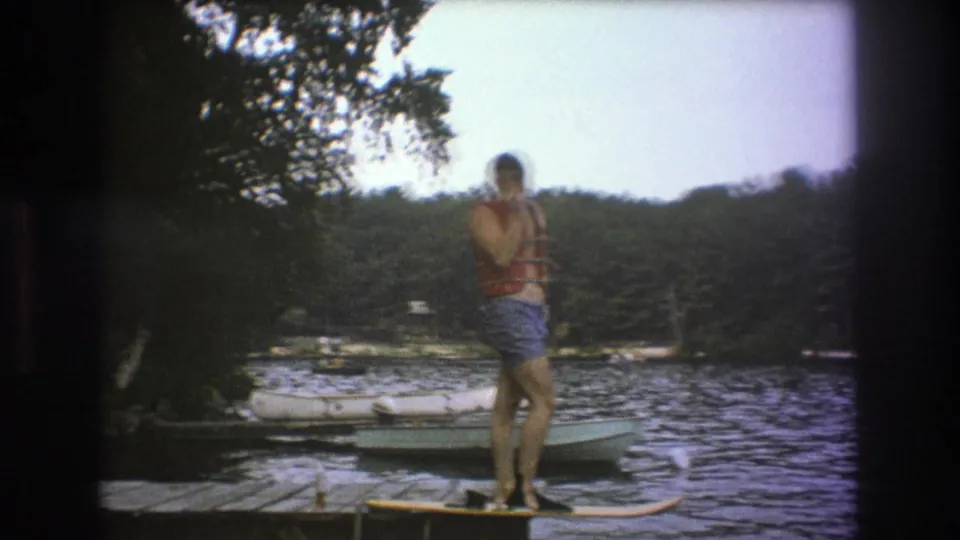 In metadata-supported 1975 Boston-area 8mm footage, a man in a red life vest and blue swim trunks stands on a wooden dock with lake water, boats, and trees behind him.