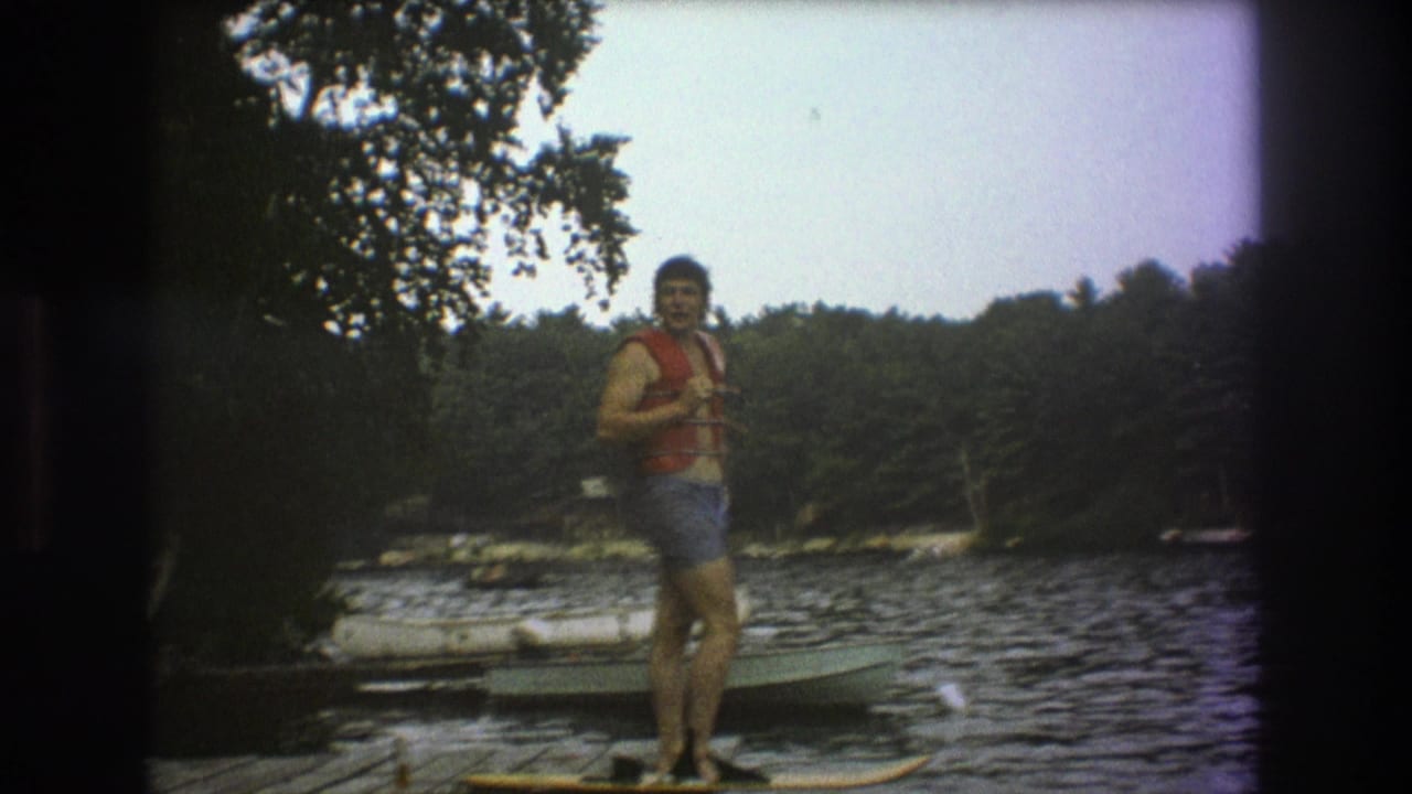 The man stands near the edge of a Boston-area lake dock, facing the camera in a red life vest as small boats sit on the water behind him.