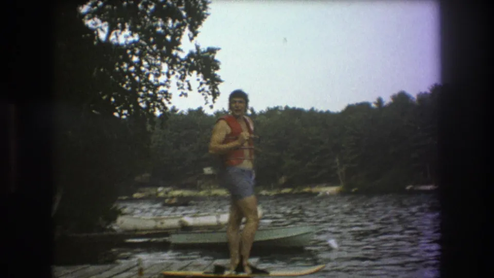 The man stands near the edge of a Boston-area lake dock, facing the camera in a red life vest as small boats sit on the water behind him.