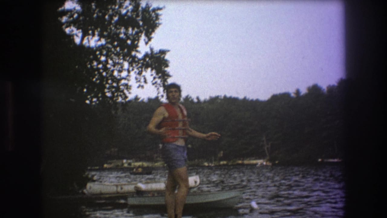A man in a red life vest gestures while standing on a wooden dock, with rippled lake water, small boats, and trees visible in the soft 8mm frame.