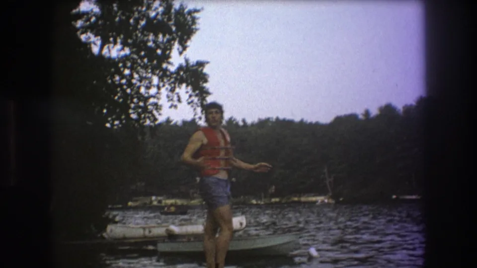 A man in a red life vest gestures while standing on a wooden dock, with rippled lake water, small boats, and trees visible in the soft 8mm frame.