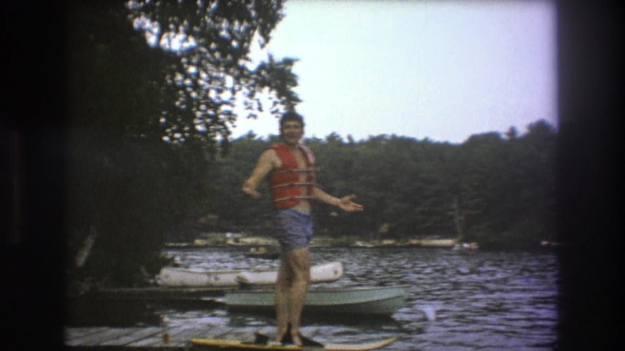 The man holds a casual pose on the lakeside dock, one hand extended toward the camera, with a board at his feet and boats near the wooded shore behind him.