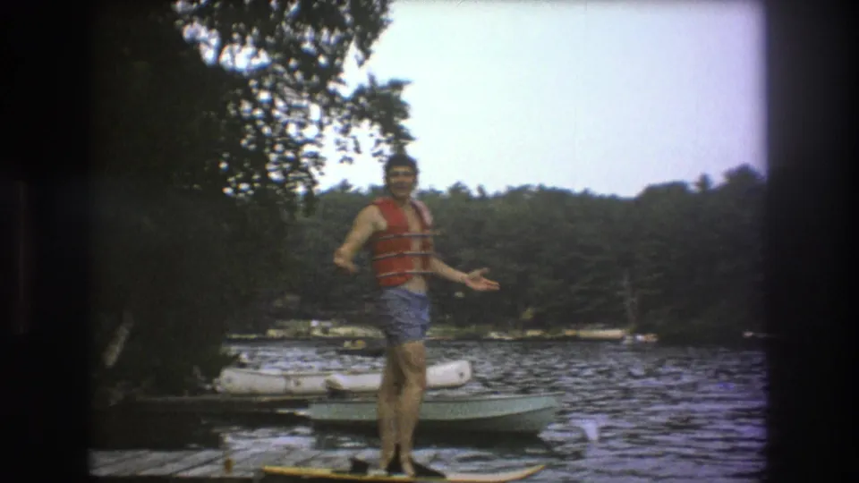 The man holds a casual pose on the lakeside dock, one hand extended toward the camera, with a board at his feet and boats near the wooded shore behind him.