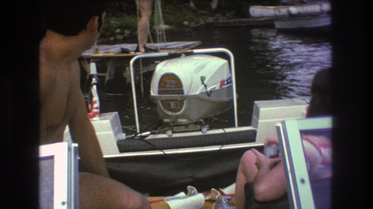 1975 Boston home-movie frame of a shirtless man and seated woman on a motorboat, with a white outboard engine and watercraft in the background.