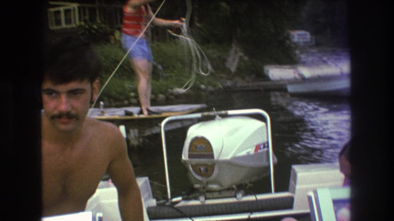 1975 Boston-area 8mm frame showing a shirtless mustached man in the foreground of a boat while another person in a red life vest stands on a dock handling rope behind him.