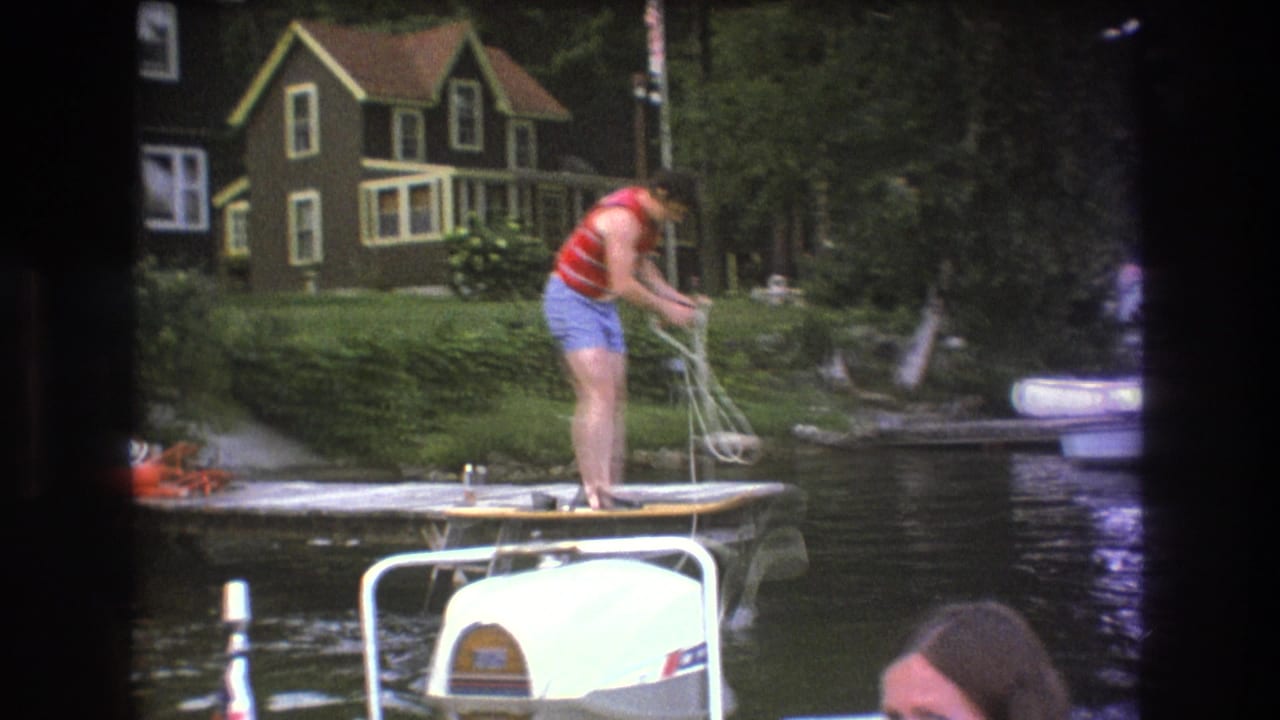 1975 Boston-area 8mm frame showing a person in a red life vest and blue shorts handling rope on a wooden dock beside lake water, with a house and boat visible in the background.