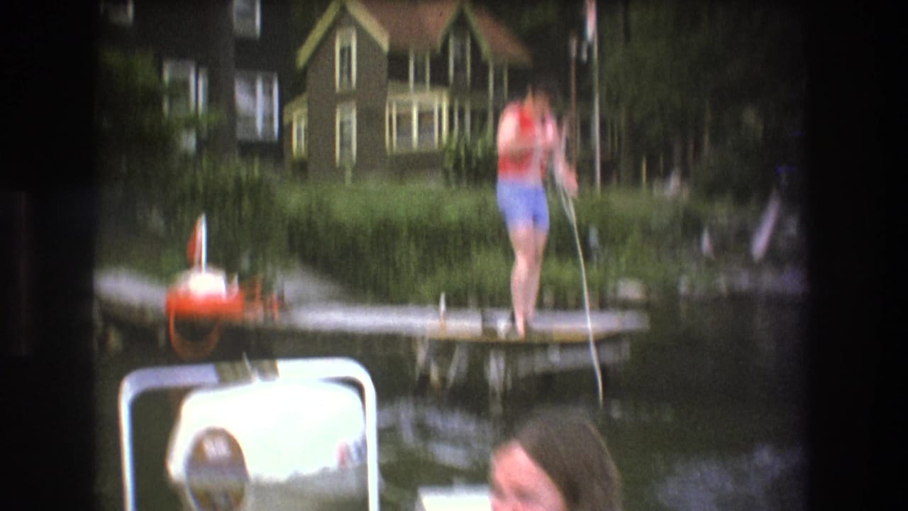 Soft-focused 1975 Boston-area 8mm frame showing a person on a wooden dock handling rope above a boat motor, with lake water and a house along the shore.