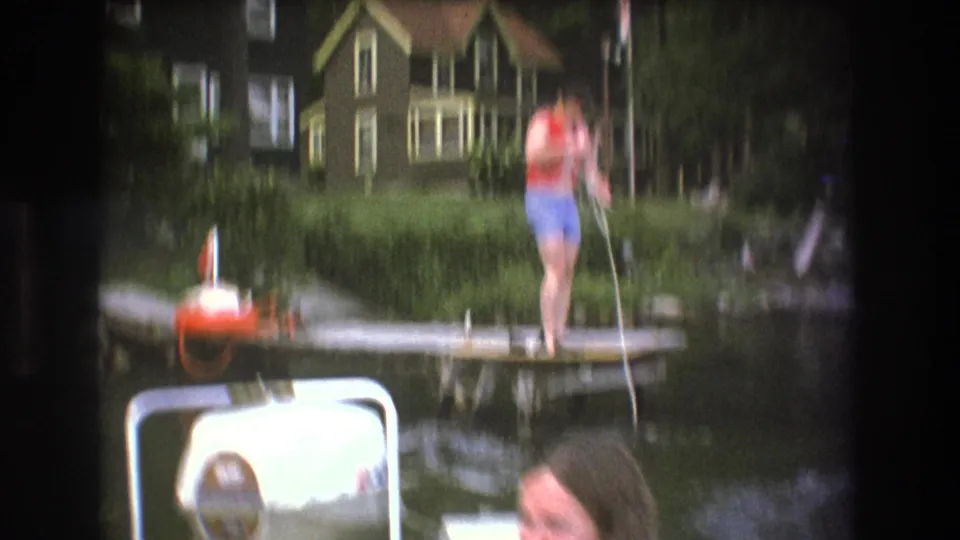 Soft-focused 1975 Boston-area 8mm frame showing a person on a wooden dock handling rope above a boat motor, with lake water and a house along the shore.