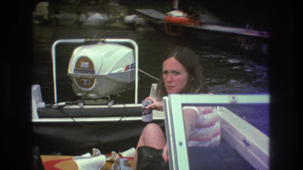 1975 Boston-area 8mm frame showing a woman in a striped sleeveless top seated in a boat near a white outboard motor, with lake water and dock equipment behind her.