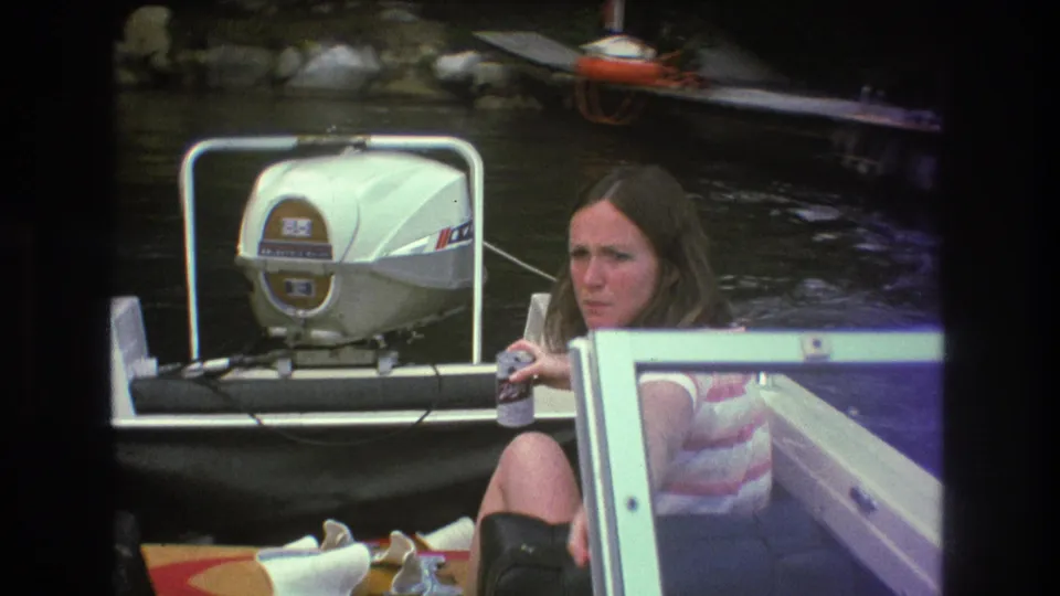 1975 Boston-area 8mm frame showing a woman in a striped sleeveless top seated in a boat near a white outboard motor, with lake water and dock equipment behind her.