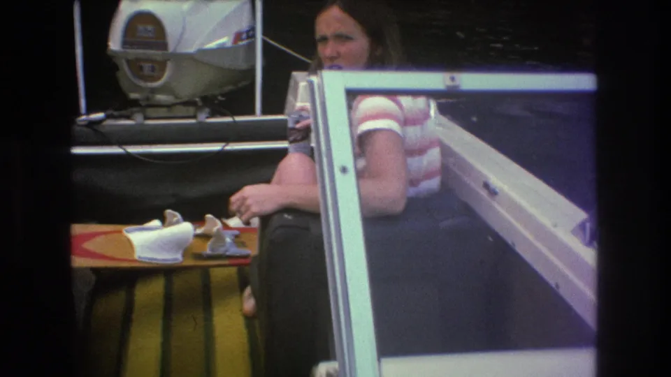 A seated woman is partly framed by a boat windshield while a white outboard motor sits behind her, matching the supplied 1975 lakeside home-movie context.