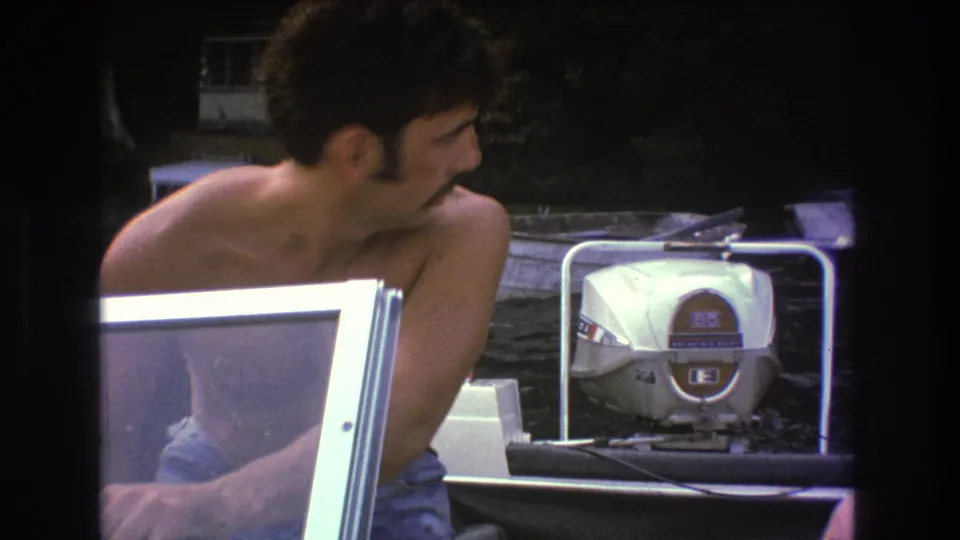 A shirtless adult man sits at the helm of a small boat, with a windshield edge in the foreground and a white outboard motor visible behind him.