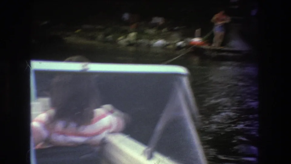 A soft, handheld Super 8 frame looks past the boat windshield toward a distant person appearing to water ski on dark lake water.