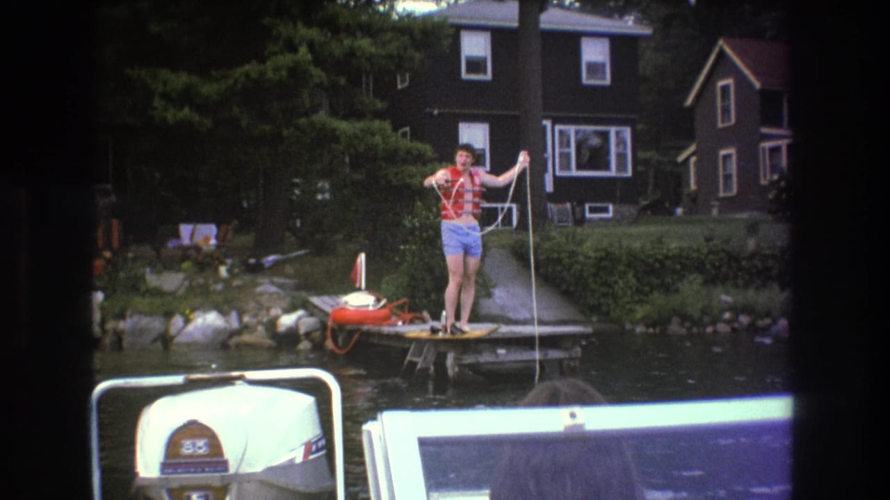 In 1975 Boston-area 8mm footage, a man in a red life vest and blue trunks stands on a wooden dock holding a rope as a white outboard motor appears in the foreground.
