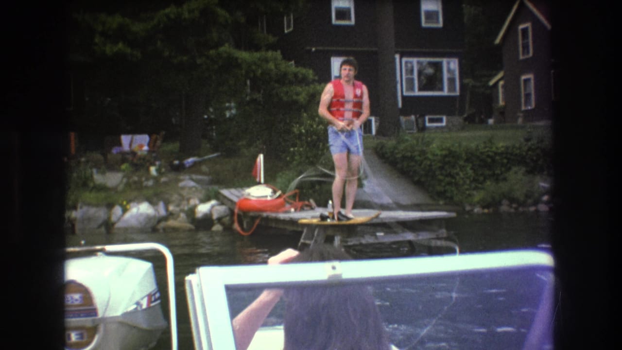 A man in a red life vest gathers the rope at the edge of a wooden dock, with water, shoreline rocks, trees, and a lakeside house visible in this 1975 Boston-area 8mm frame.