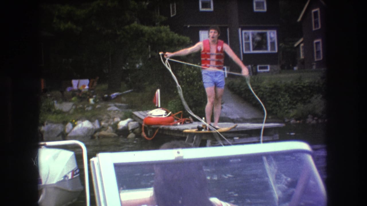 The man extends the rope across the dock as if preparing to secure a boat, with a white outboard motor, water, trees, rocks, and a dark lakeside house in view.