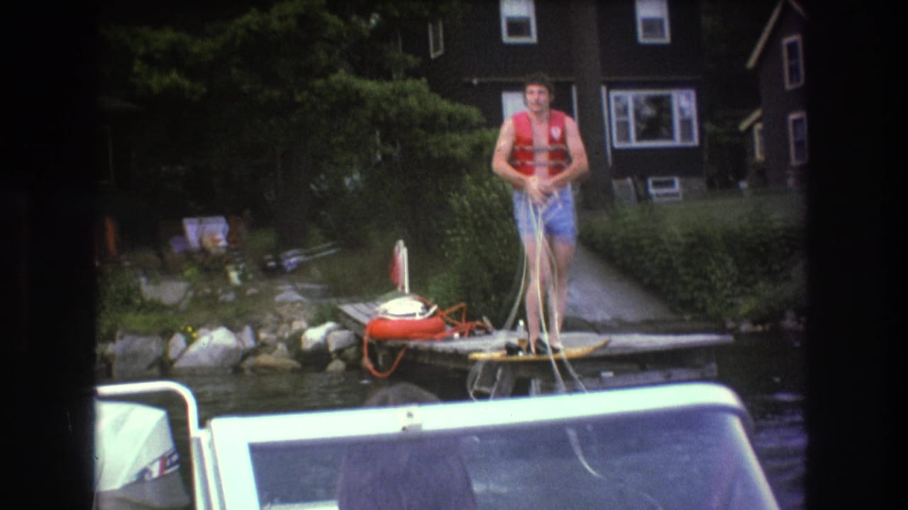 In 1975 Boston-area home-movie footage, a man in a red life jacket and blue swim trunks stands on a wooden dock with a tow rope as a motorboat fills the foreground.