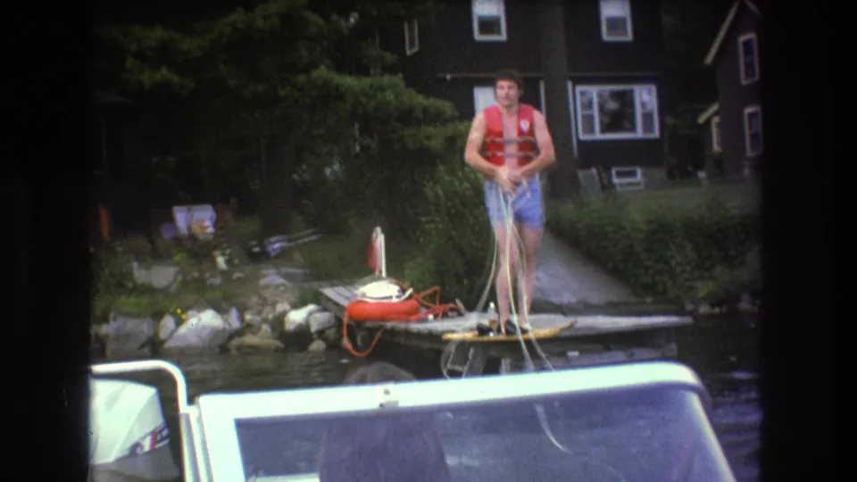 In 1975 Boston-area home-movie footage, a man in a red life jacket and blue swim trunks stands on a wooden dock with a tow rope as a motorboat fills the foreground.